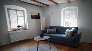 A living room with a blue sofa, a small round table, a radiator, and a window with a view. - Apartment 'La Maison Palatinate' with Charm, Southern Wine Route (Kapsweyer)