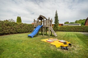 Una estructura de juego de madera con un tobogán y una escalera de cuerda, una manta amarilla con juguetes, y un excavador de juguete sobre un césped.