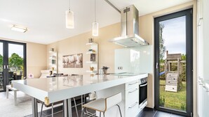 Une cuisine moderne dotée d’un grand îlot central, d’appareils électroménagers en acier inoxydable et d’une porte-fenêtre coulissante donnant sur l’extérieur.