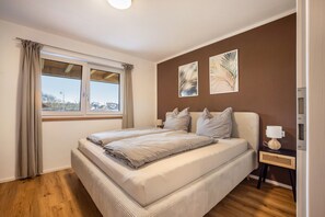 Een slaapkamer met een groot bed, een raam met gordijnen, een nachtkastje met een lamp, en twee kunstwerken aan de muur.