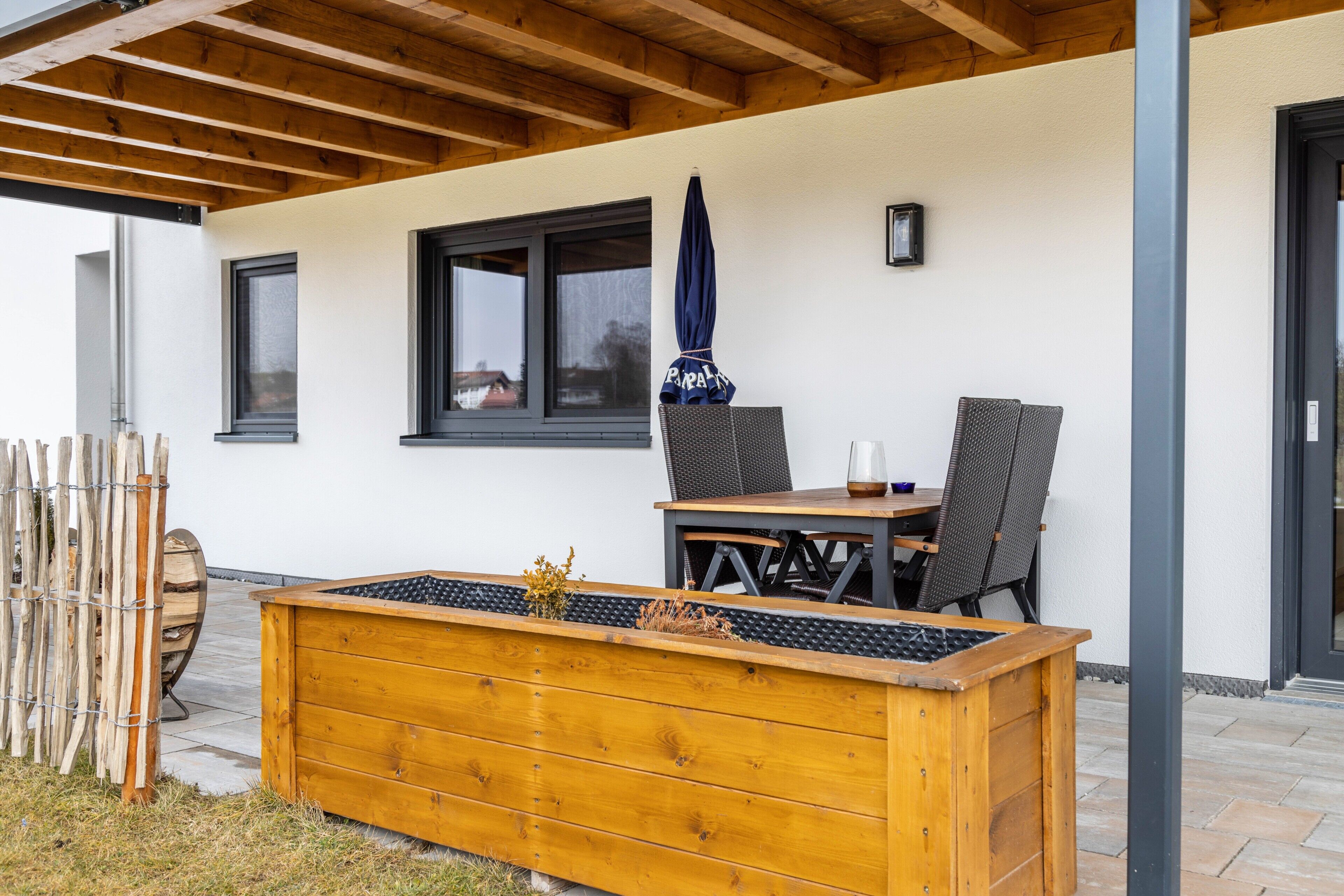 A covered patio with a wooden table and chairs, a large planter box, and a window with a view.