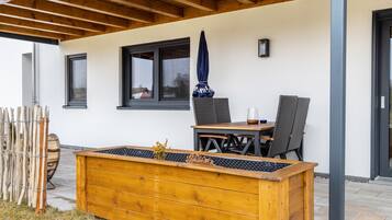 A covered patio with a wooden table and chairs, a large planter box, and a window with a view.