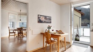 Un coin repas avec des meubles en bois, une table agrémentée d’un vase et de verres à vin, et une vue sur un paysage montagneux par une porte vitrée.