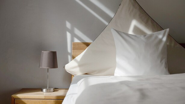 A neatly made bed with white linens, a bedside lamp, and a pillow against a wall with a sloped ceiling.