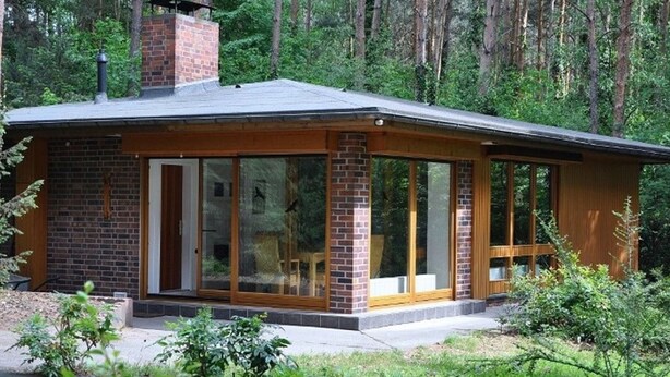 Eine eingeschossige Holzhütte mit einem Backsteinsschornstein, großen Schiebegläsern und einer überdachten Veranda.