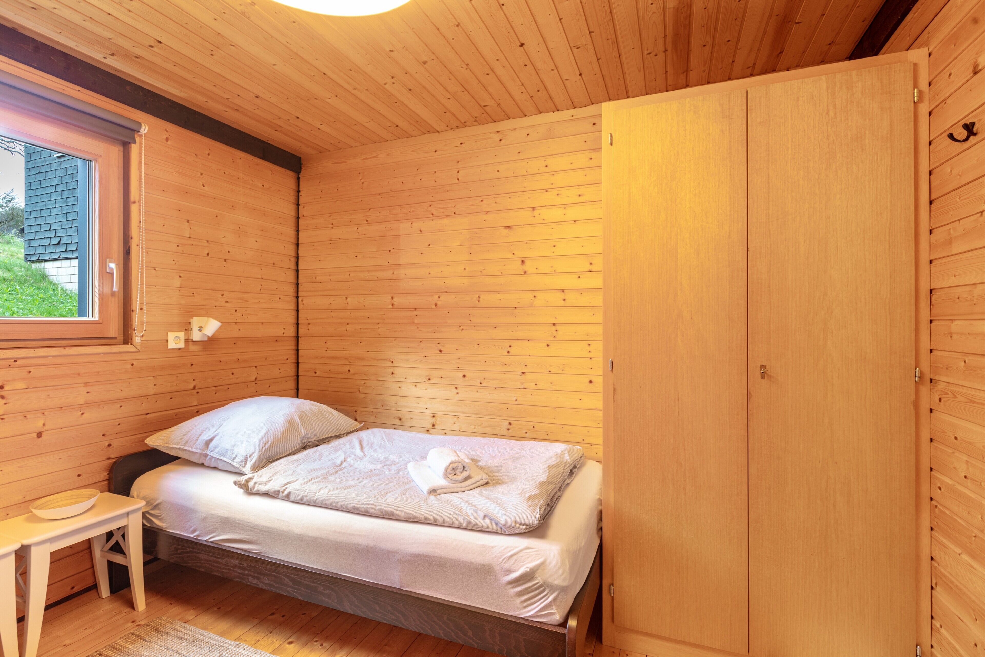 A wooden room with a bed, a small table, and a window.