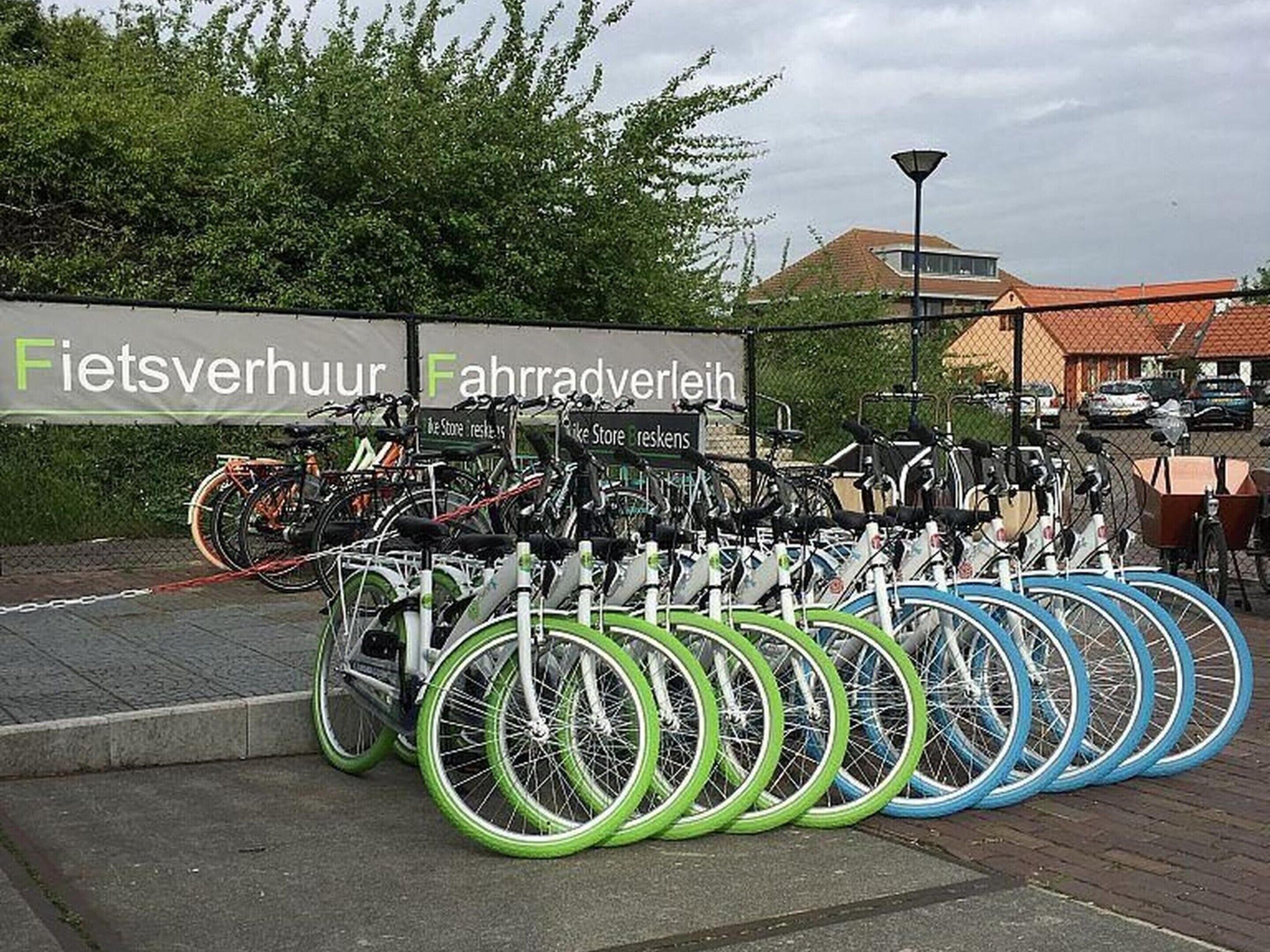 Zona de estacionamiento para bicicletas con varias bicicletas y un letrero que dice 'Fietsverhuur Fahrradverleih'.