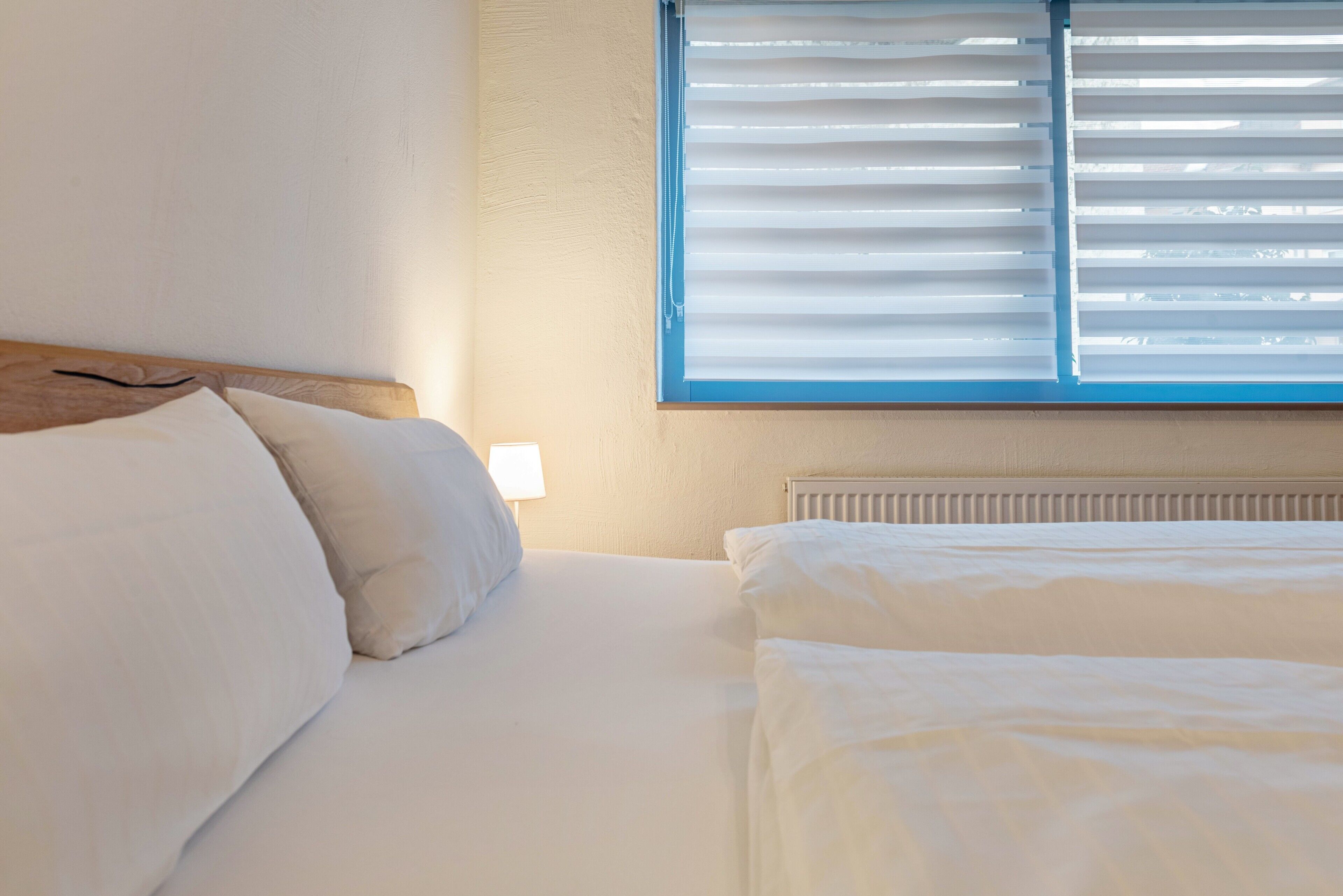 Una habitación de hotel con una cama, una ventana con persianas, una lámpara de noche y un radiador.