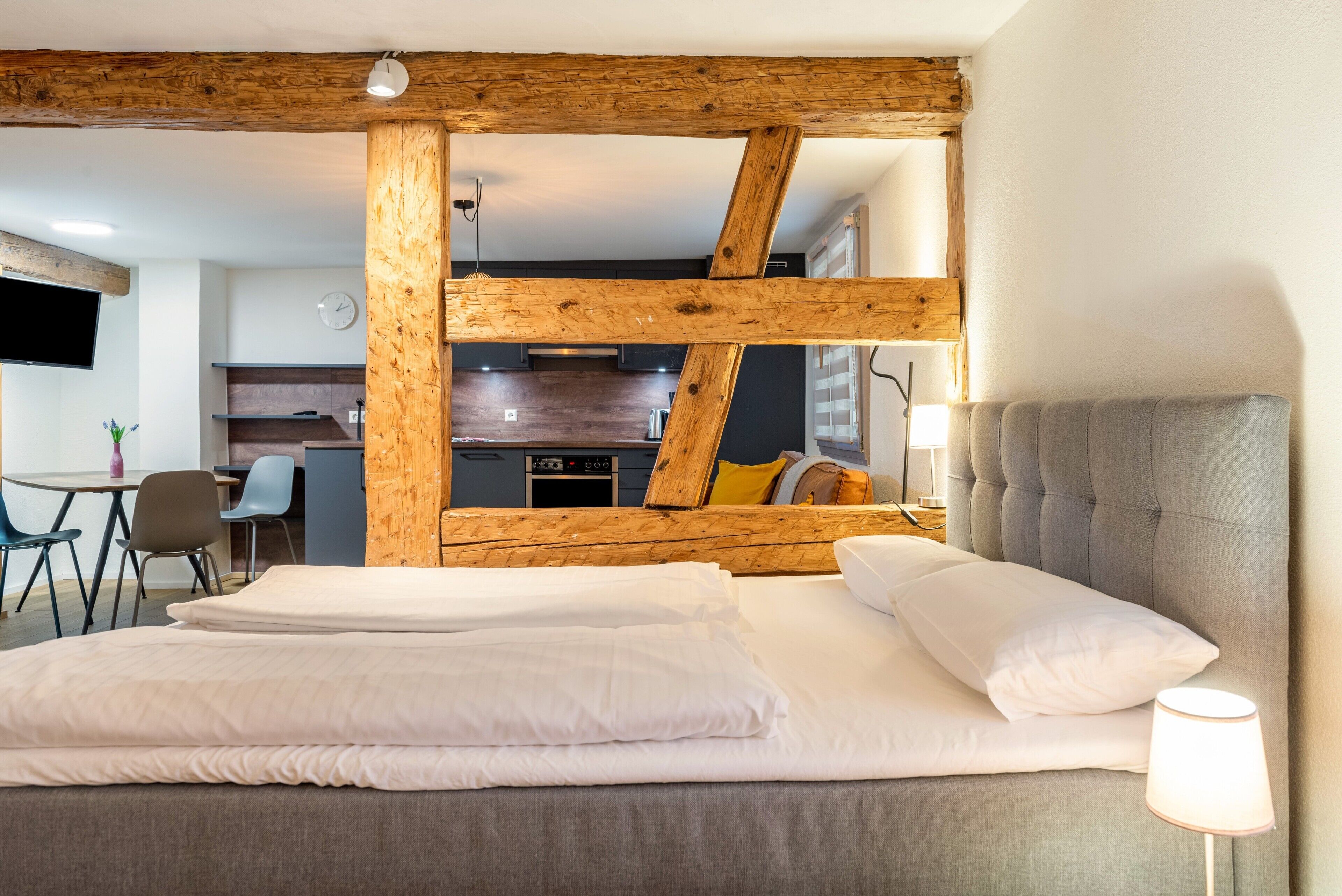A bedroom with a bed, a lamp, and a view of a kitchen and dining area.