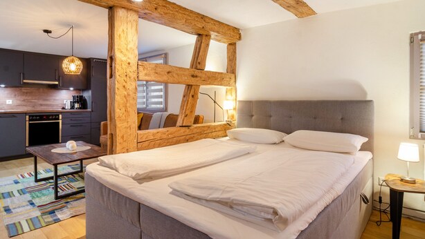 A bedroom with a large bed, a wooden ceiling beam, a bedside table with a lamp, and a view of the kitchen. - Apartment '3' in Bodman-Ludwigshafen (Bodman-Ludwigshafen)