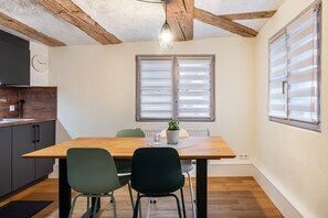 Uma sala de jantar com uma mesa de madeira, duas cadeiras, uma planta em vaso e uma janela com persianas.