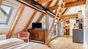 Un dormitorio con zona de descanso elevada, vigas de madera, un televisor, una silla rosa y un aparador de madera.