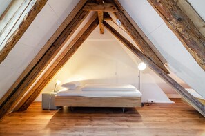 A small, cozy attic bedroom with a wooden ceiling, a single bed, bedside lamps, and a nightstand.