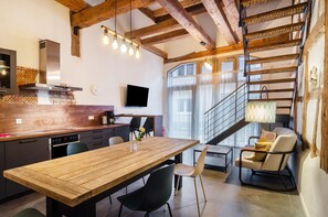 Una cocina moderna con una mesa de madera, sillas negras, un televisor y una escalera que lleva a un balcón.