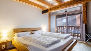 A bedroom with a wooden headboard, two beds, a nightstand with a lamp, and a balcony with a view of a building. - 'Family Apartment 6' (Bodman-Ludwigshafen)