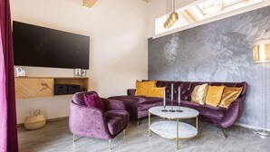 Una sala de estar moderna con un televisor de pantalla plana grande, un sofá morado y una mesa de centro redonda, además de un mural decorativo en la pared.