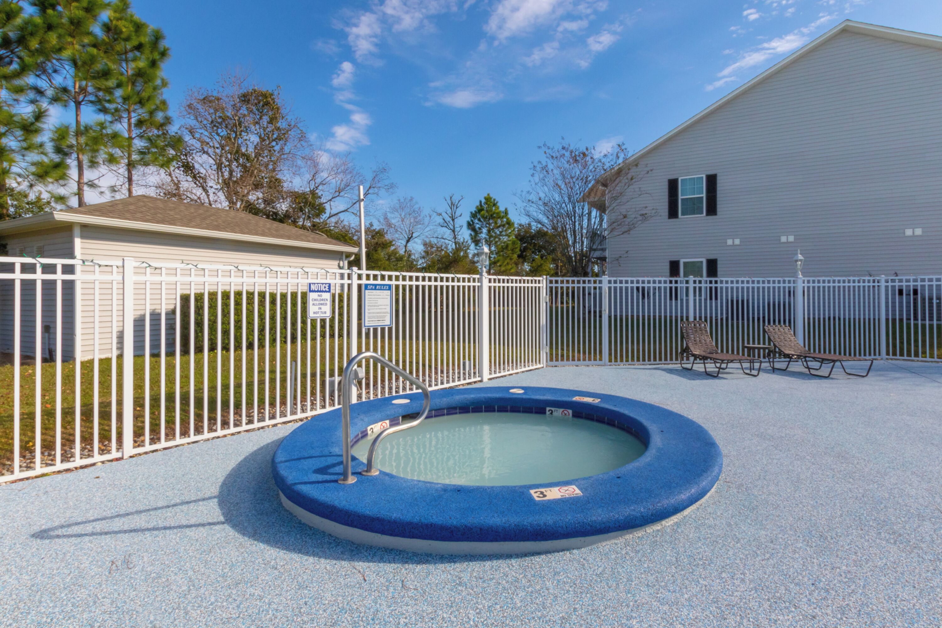 A swimming pool with a white fence, a lounge area with two chairs, and a building in the background.