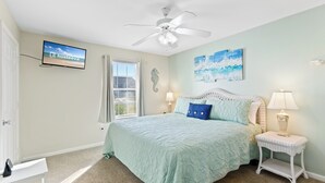 Ein Schlafzimmer mit Bett, Deckenventilator, an der Wand montiertem Fernseher, Fenster und Nachttisch mit Lampe.