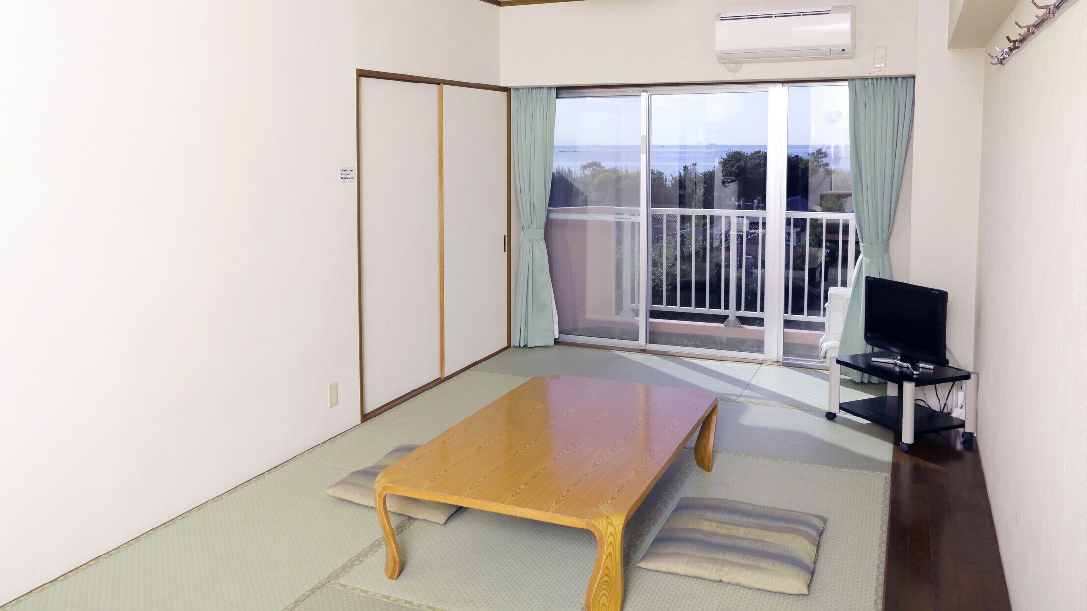 A room with a sliding glass door to a balcony, a wooden table, a black TV stand, and a tatami mat floor.