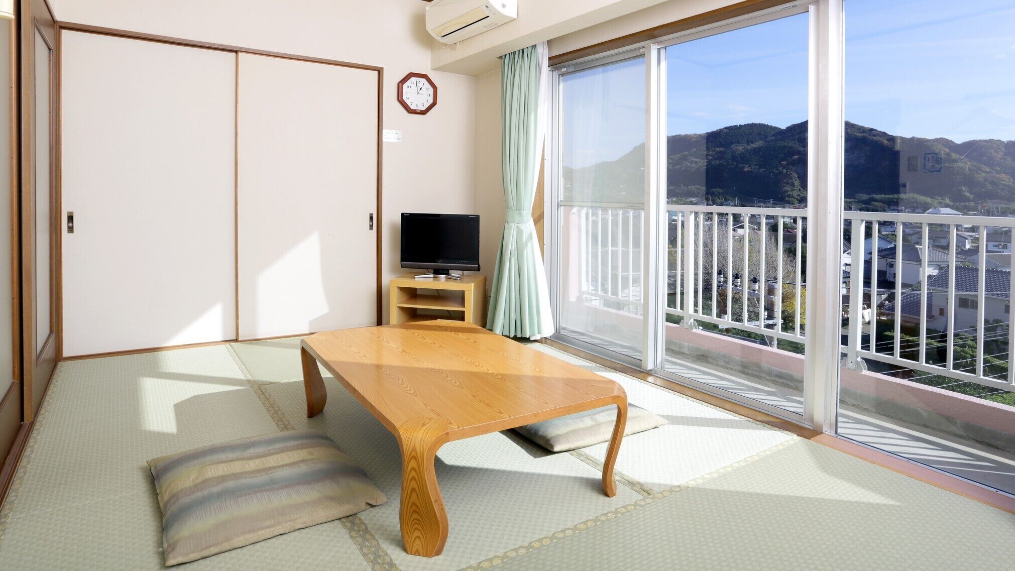 A traditional Japanese room with tatami flooring, a wooden table, a television, and a sliding door leading to a balcony with a view of hills.