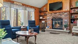 Una sala de estar acogedora con chimenea, dos sillones azules, una mesa de centro y un televisor montado en la pared.