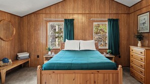 Un dormitorio con paneles de madera, una cama, dos ventanas con cortinas, un banco, un aparador y un espejo redondo montado en la pared.