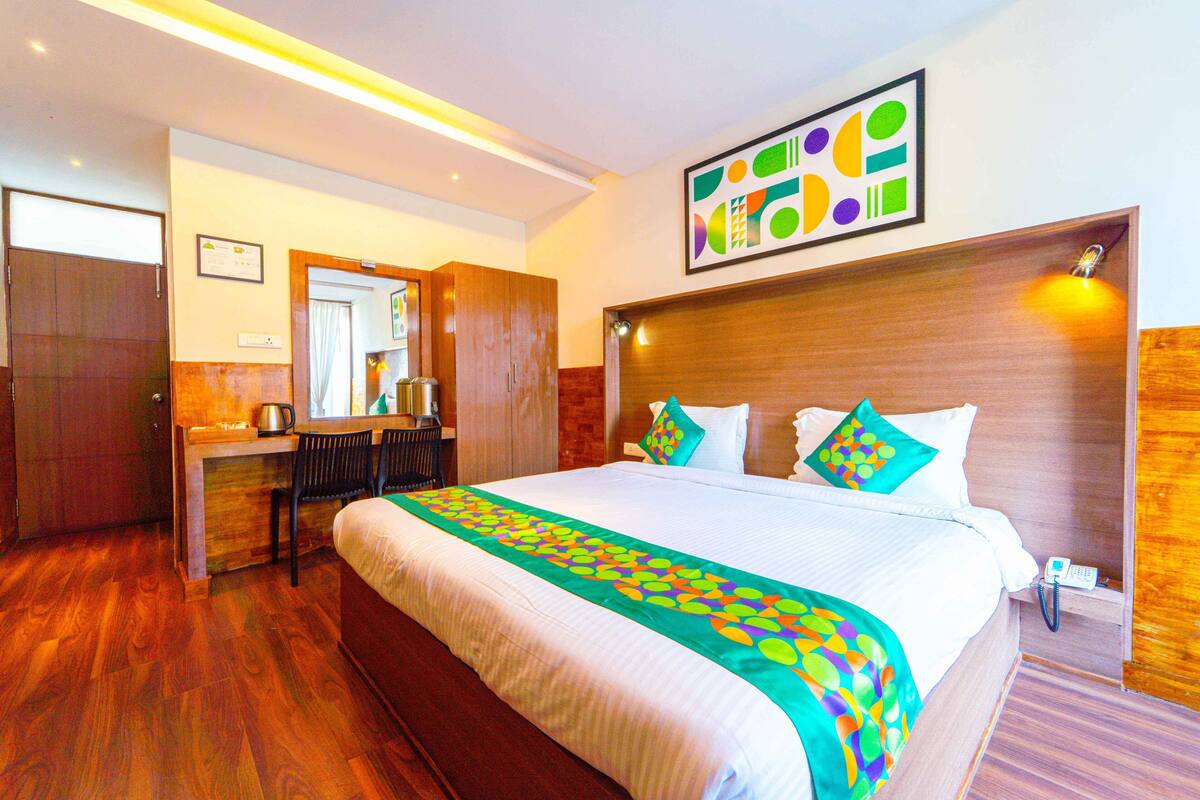 Quarto de hotel com uma cama grande, uma escrivaninha com cadeira, um espelho e cabeceira de madeira.