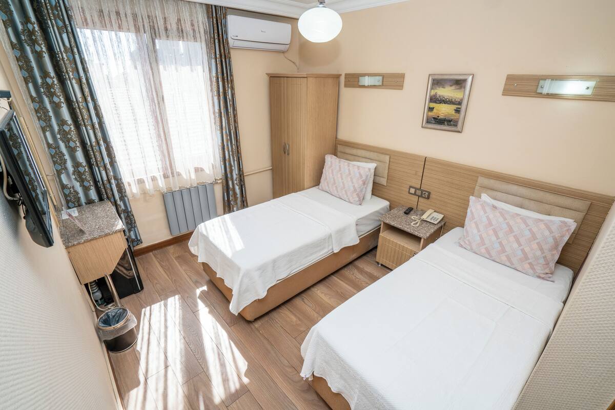 A hotel room with two beds, a wooden wardrobe, a small bedside table with a lamp, and a window with curtains.