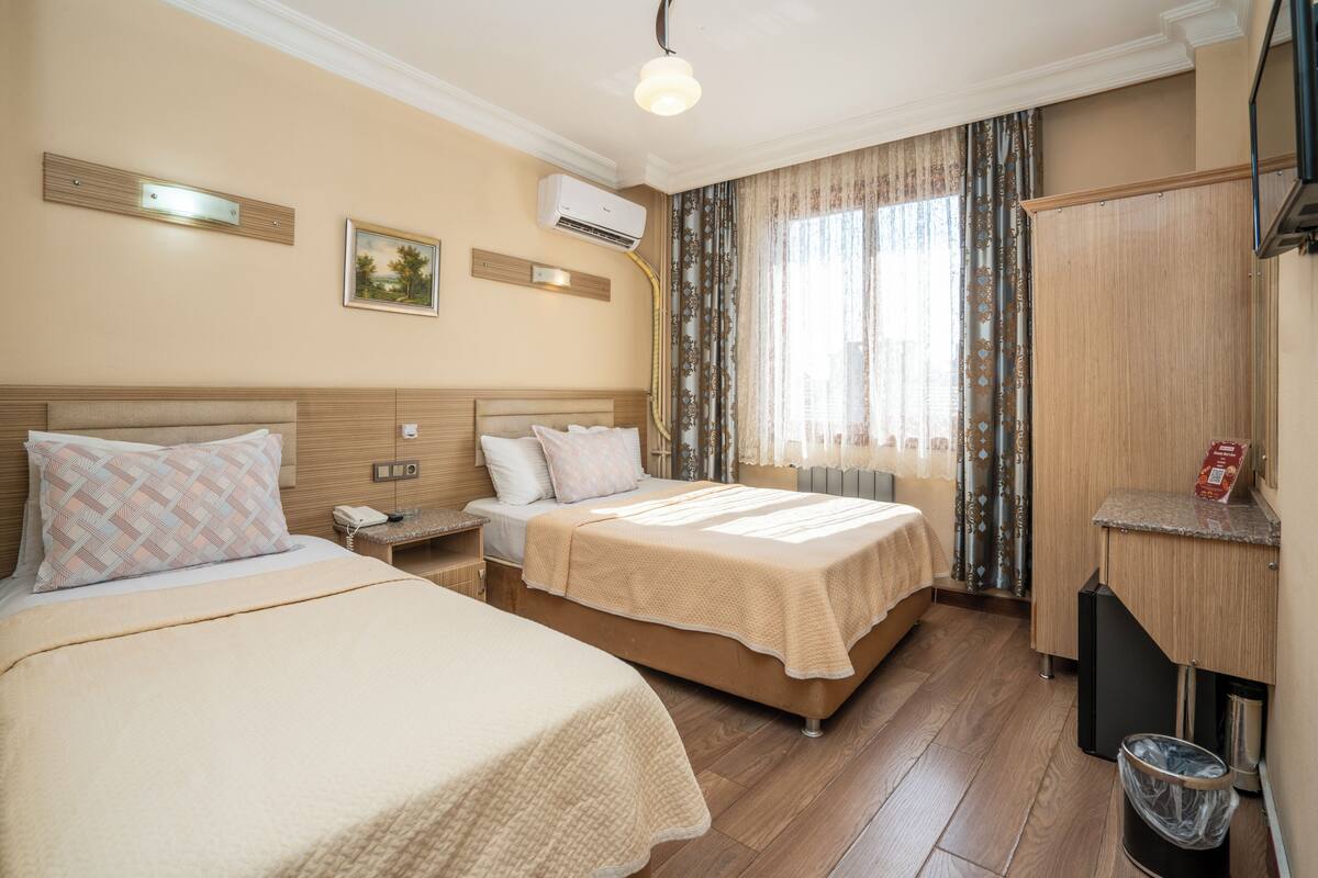 A hotel room with two beds, a wooden headboard, a bedside table with a lamp, a wall-mounted air conditioner, and a window with curtains.