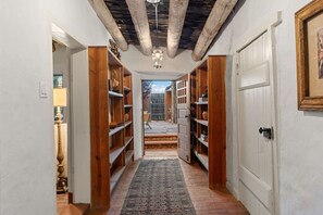 A narrow hallway with wooden bookshelves, a patterned rug, and a ceiling with exposed beams. - Ski Discounts! Downtown Historic Retreat w/ Casita (Santa Fe)