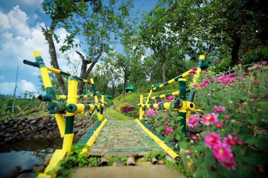 Un sendero atraviesa un jardín con estructuras amarillas y verdes, rodeado de árboles y flores.