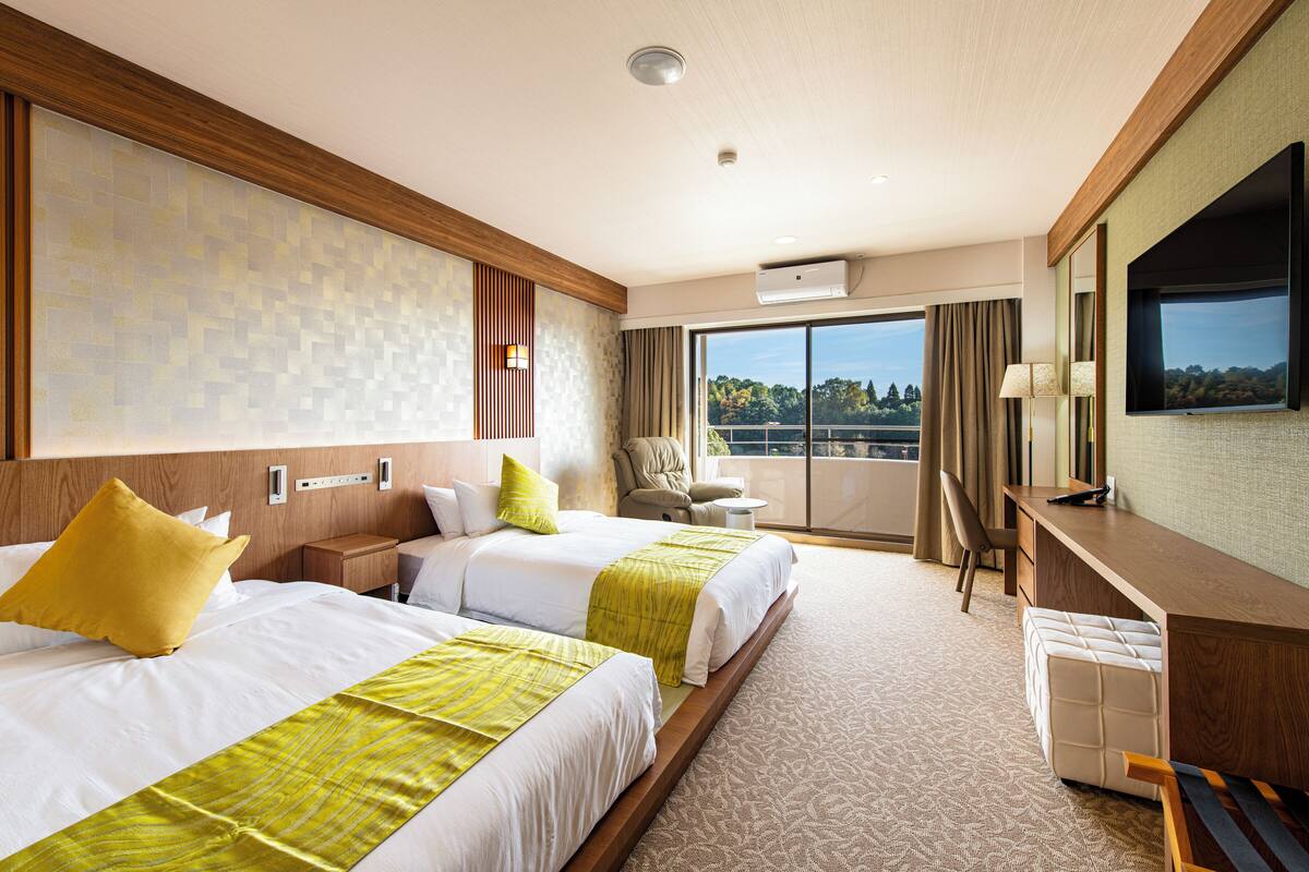 A hotel room with two beds, a desk, a chair, and a large window offering a view.