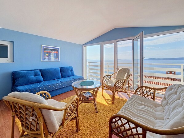 A balcony with a view of the sea, a blue sofa, wicker chairs, and a white sofa.