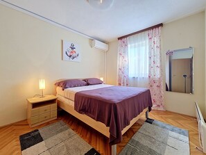 Un dormitorio con cama, mesita de noche, lámpara, unidad de aire acondicionado y ventana con cortinas.