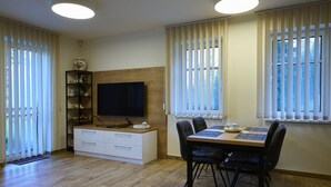 A modern living room with a wooden floor, a flat-screen TV mounted on a wooden wall, a dining table with chairs, and a bookshelf with decorative items. - Markéta by Interhome (Smržovka)