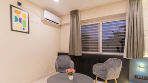 Une pièce comprenant une table ronde, deux chaises, un tableau encadré, un appareil de climatisation, une fenêtre avec des rideaux et un luminaire fixé au mur.