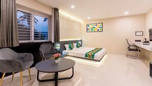 Une chambre d’hôtel moderne comprenant un lit, deux fauteuils gris, une petite table ronde avec un vase de fleurs, un bureau avec une chaise et une fenêtre avec des stores.