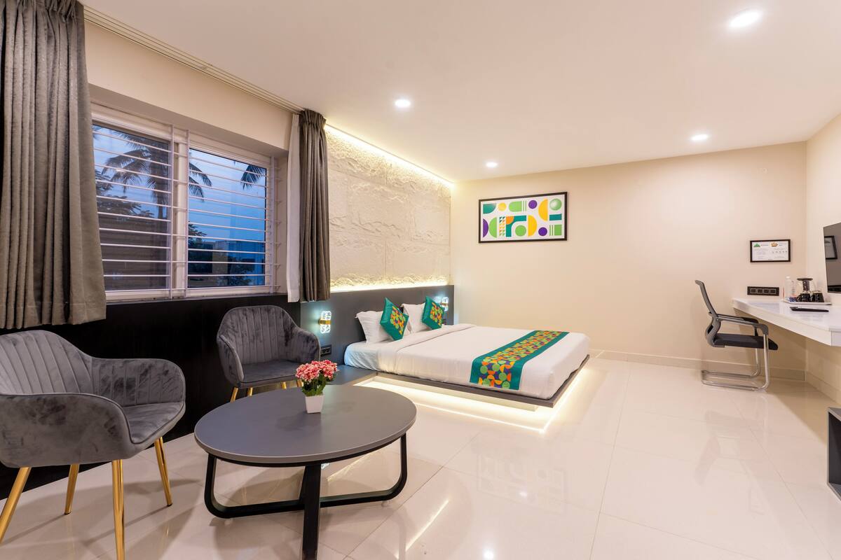 A modern hotel room with a bed, two grey armchairs, a small round table with a vase of flowers, a desk with a chair, and a window with blinds.