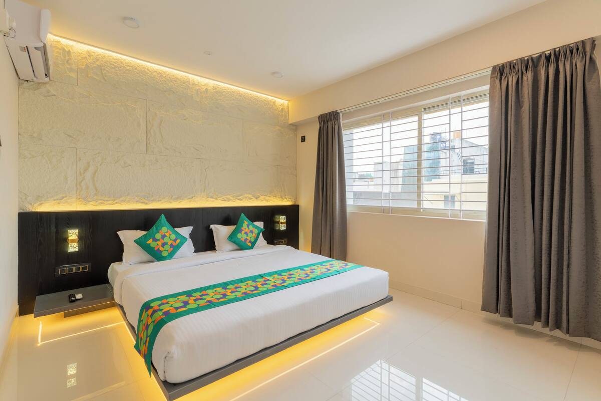 A modern hotel room with a large bed, a nightstand, a wall-mounted air conditioner, and a window with curtains.