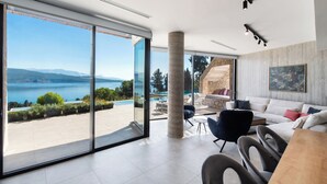 A modern living room with a large glass door leading to a pool area, a white sofa, and a dining table. - Ionian Grand Villas - Villa Naya (QPM9+9JR, Lefkada)