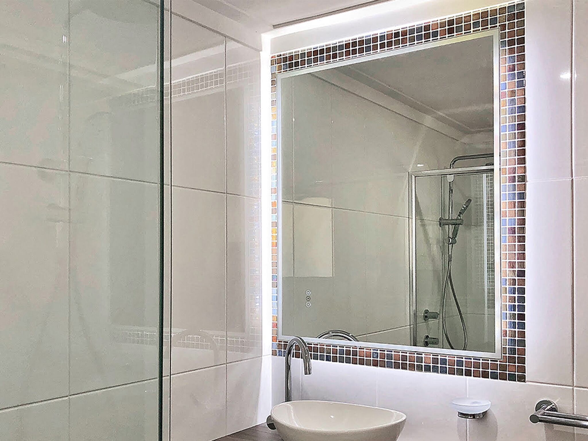 A modern bathroom with a glass shower enclosure, a large mirror with a mosaic tile border, and a sink with a chrome faucet.