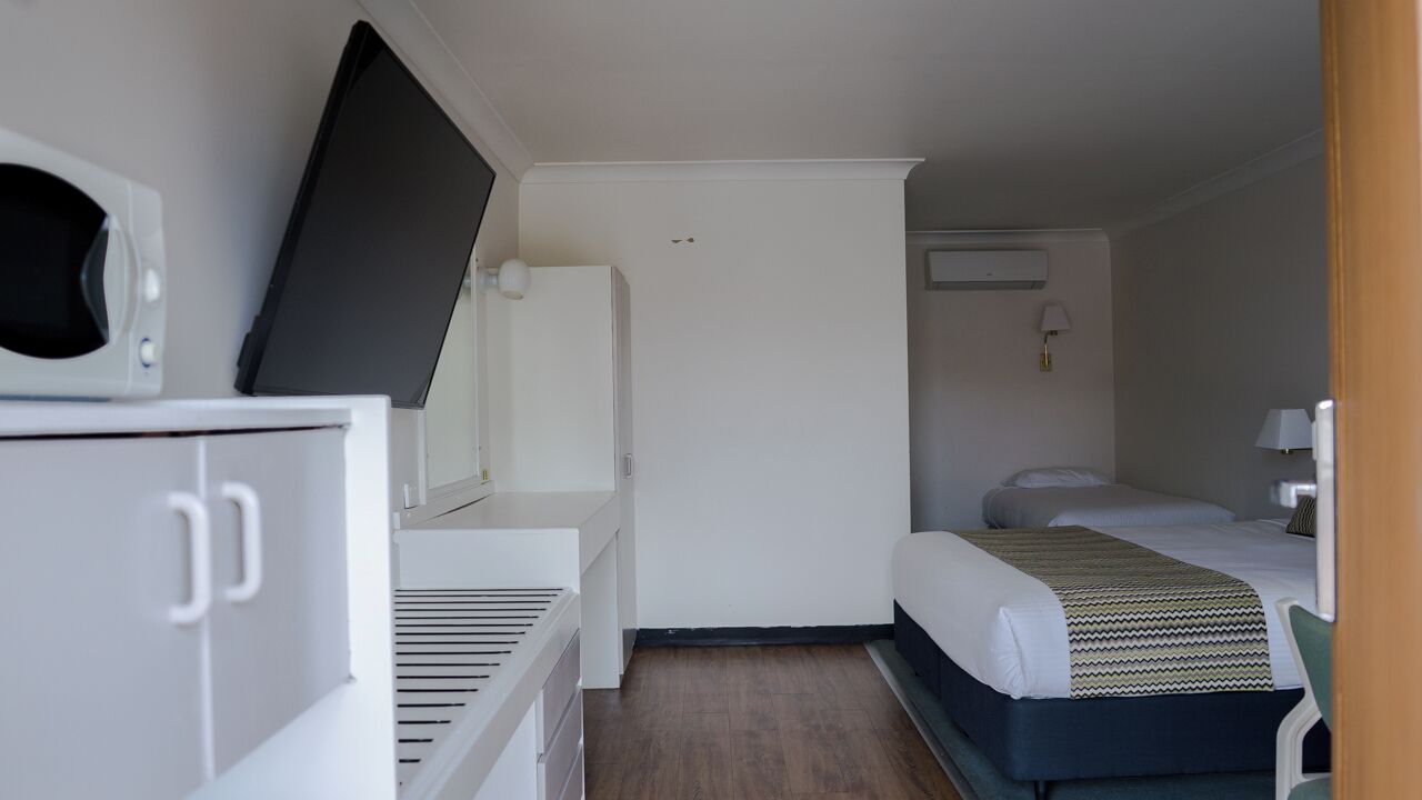 A compact hotel room with a bed, a desk with a computer, a microwave, and a small kitchenette.