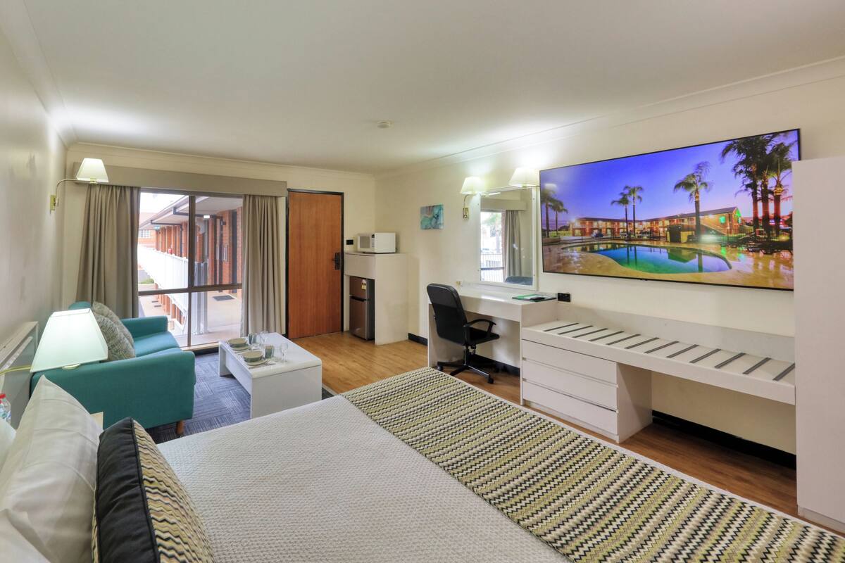A hotel room with a bed, a desk, a chair, a TV, and a balcony with a view of the pool.