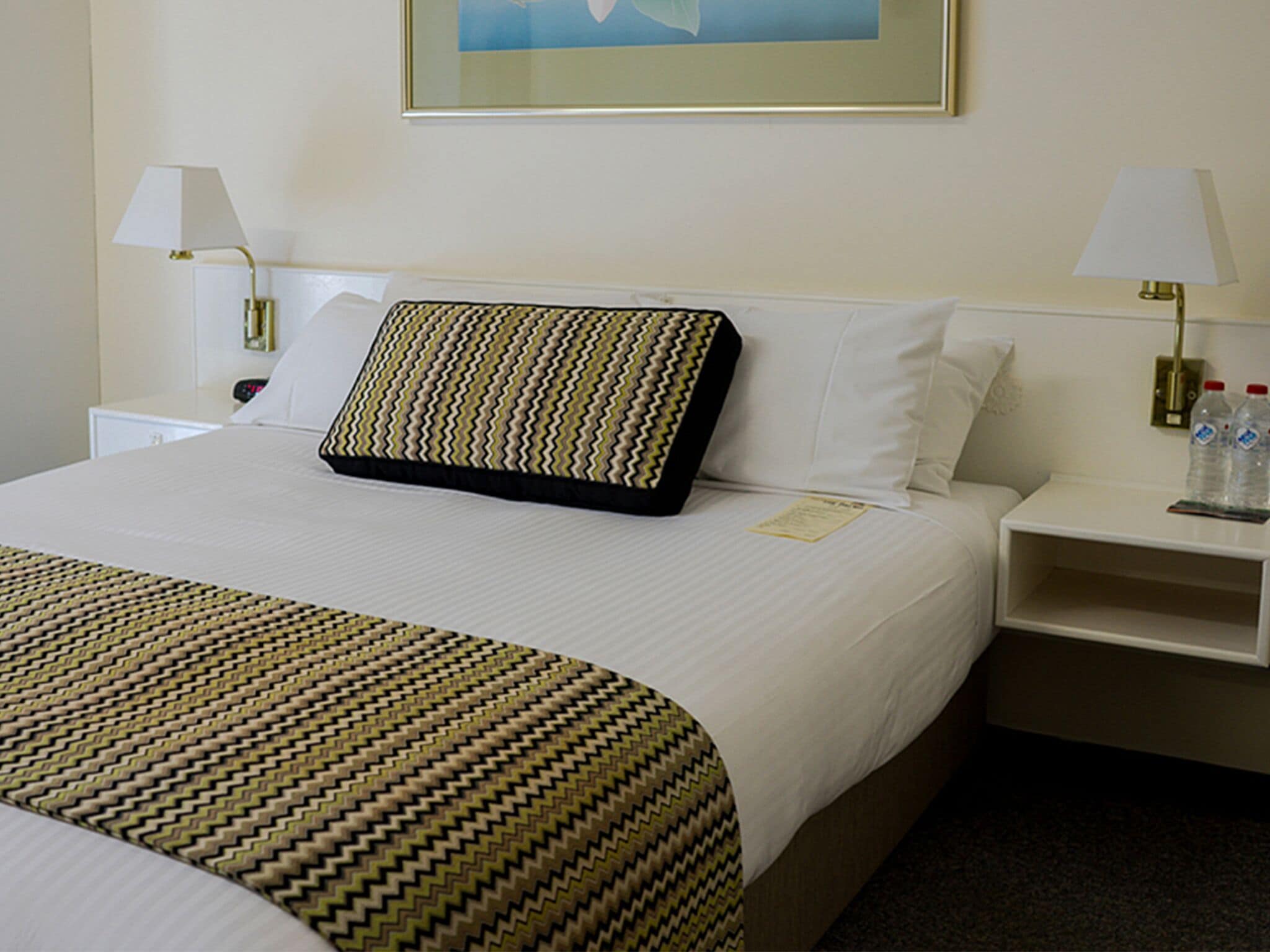 Habitación de hotel con una cama bien hecha, dos mesitas de noche con lámparas, un cuadro en la pared y una botella de agua en la mesita.