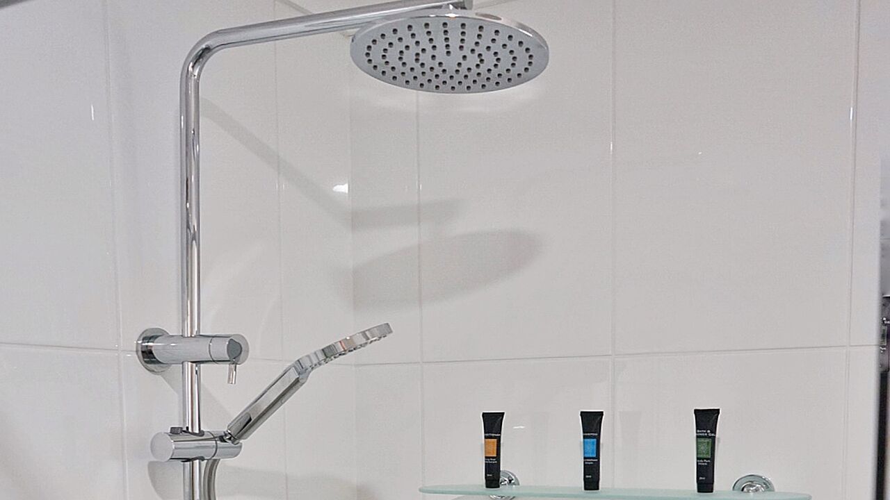 A shower with a handheld showerhead and a rain shower head. There are three toiletries on a glass shelf below.