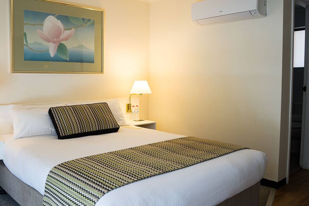 A hotel room with a large bed, two bedside lamps, a wall-mounted air conditioner, and a framed floral painting.