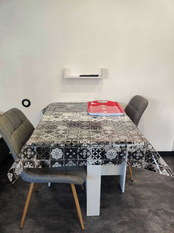 Uma mesa de jantar com toalha de padrão, duas cadeiras estofadas e uma parede branca com uma prateleira acima.