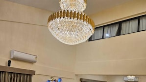 A spacious living room with a large sectional sofa, a coffee table, and a chandelier. There is a dining area with a table and chairs in the background. - Shiv Tirth By 97 Royal Vista  (Mawal)