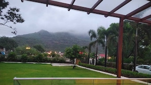 A view from a balcony with a glass railing overlooking a grassy area, a mountain in the background, and a few buildings with lights. - Shiv Tirth By 97 Royal Vista  (Mawal)