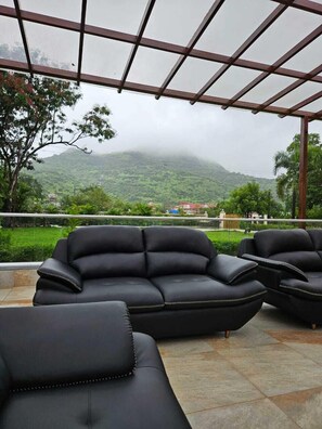 A covered outdoor seating area with black leather sofas and a view of a mountainous landscape. - Shiv Tirth By 97 Royal Vista  (Mawal)